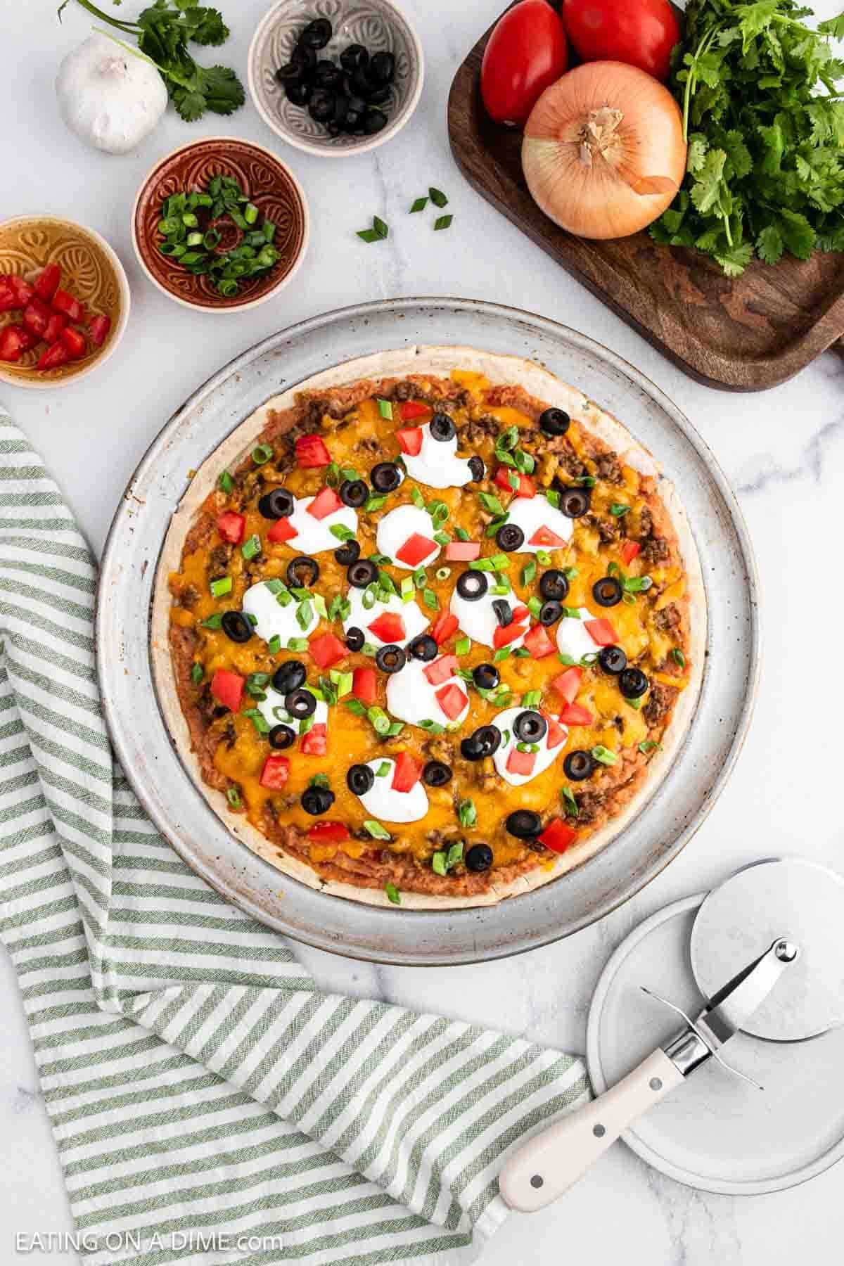 A delicious taco pizza recipe topped with melted cheese, tomatoes, olives, green onions, and sour cream sits on a round tray. Bowls of toppings, fresh veggies, and a pizza cutter rest nearby on a striped towel.