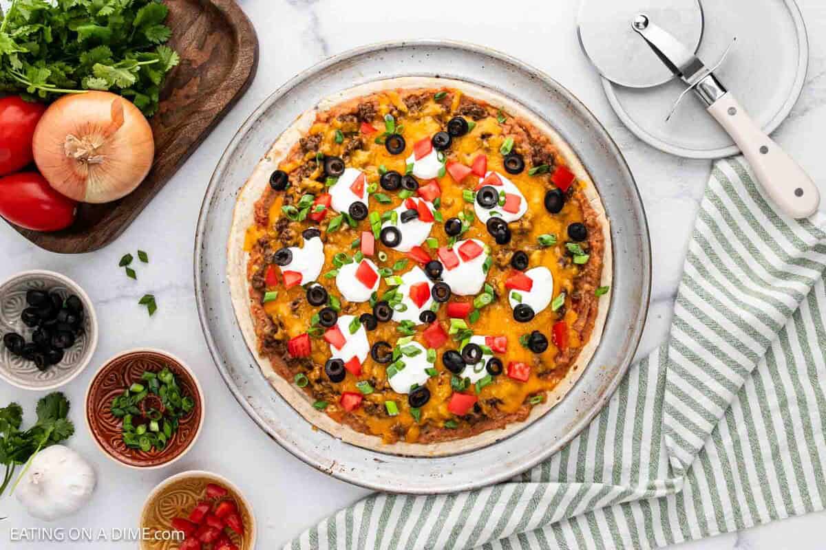 A homemade taco pizza recipe topped with cheese, olives, tomatoes, green onions, and dollops of sour cream on a white plate, surrounded by fresh ingredients and a pizza cutter on a striped towel.