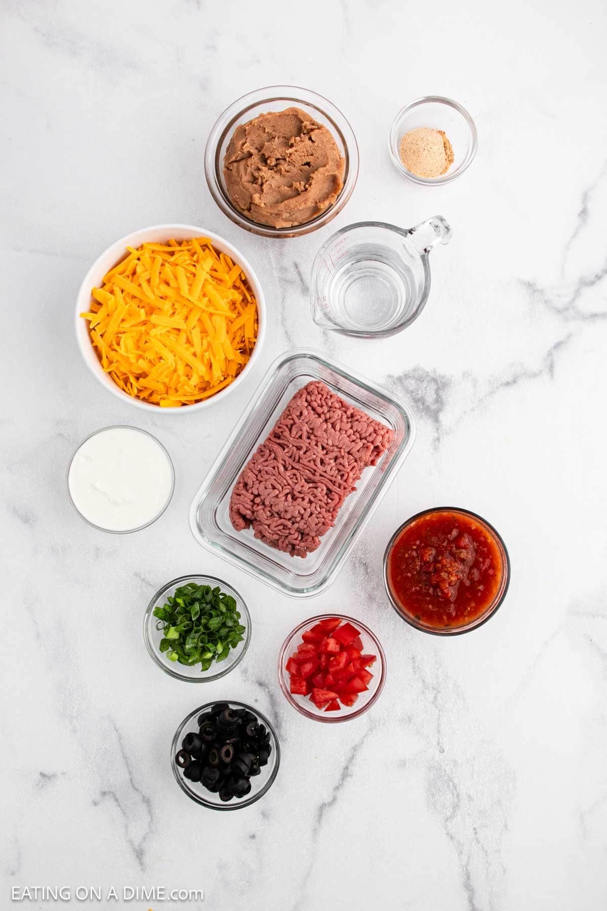 Various taco dip ingredients are arranged on a white marble surface, perfect for inspiring your next taco pizza recipe—featuring shredded cheddar cheese, refried beans, ground beef, salsa, chopped tomatoes, black olives, and green onions.