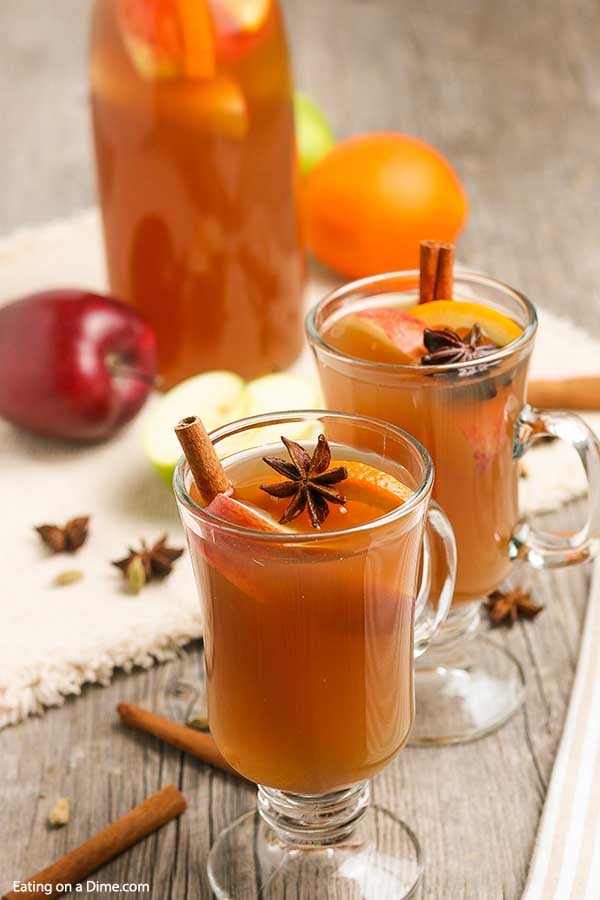 Apple Cider in clear mugs topped with cinnamon sticks and fresh apples and oranges