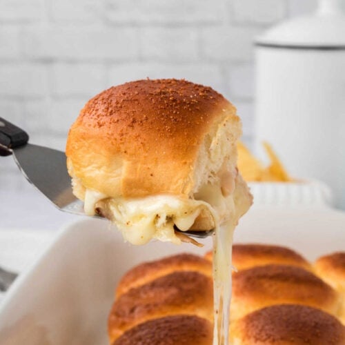 A spatula lifts a cheesy Turkey and Cheese Slider with a golden-brown top from a dish, melted cheese stretching from the sandwich.