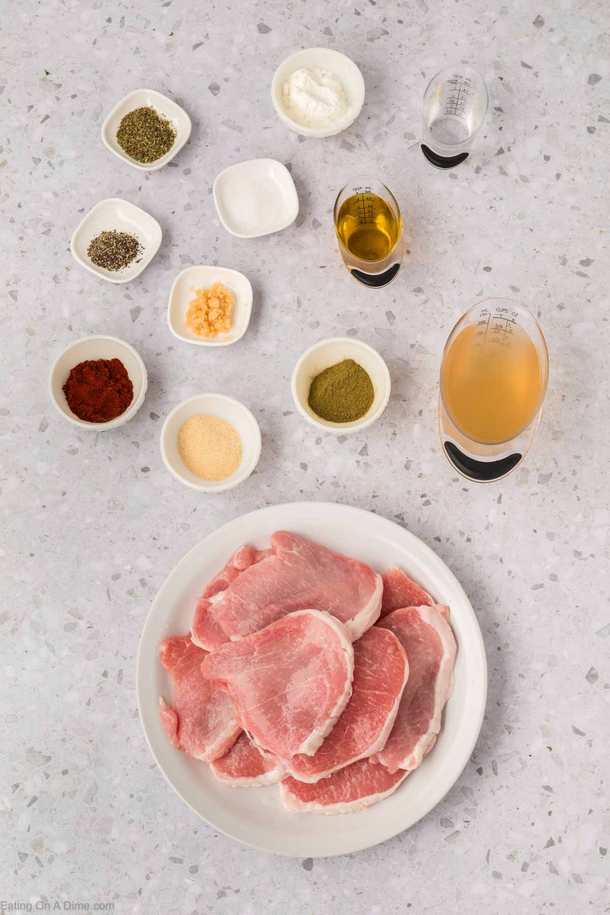 A white plate with raw pork chops on a counter, surrounded by small bowls of spices, oil, minced garlic, and two measuring cups with liquids—perfect for preparing Instant Pot Pork Chops with Gravy.