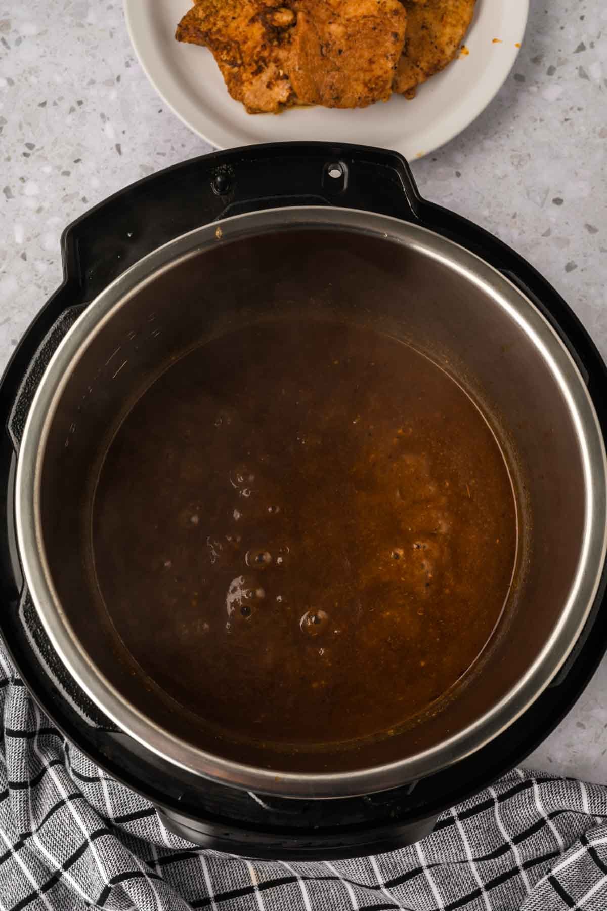 An Instant Pot Pork Chops with Gravy dish bubbles with rich brown sauce on a gray countertop, beside a black and white checkered towel and a white plate holding tender cooked chicken pieces.