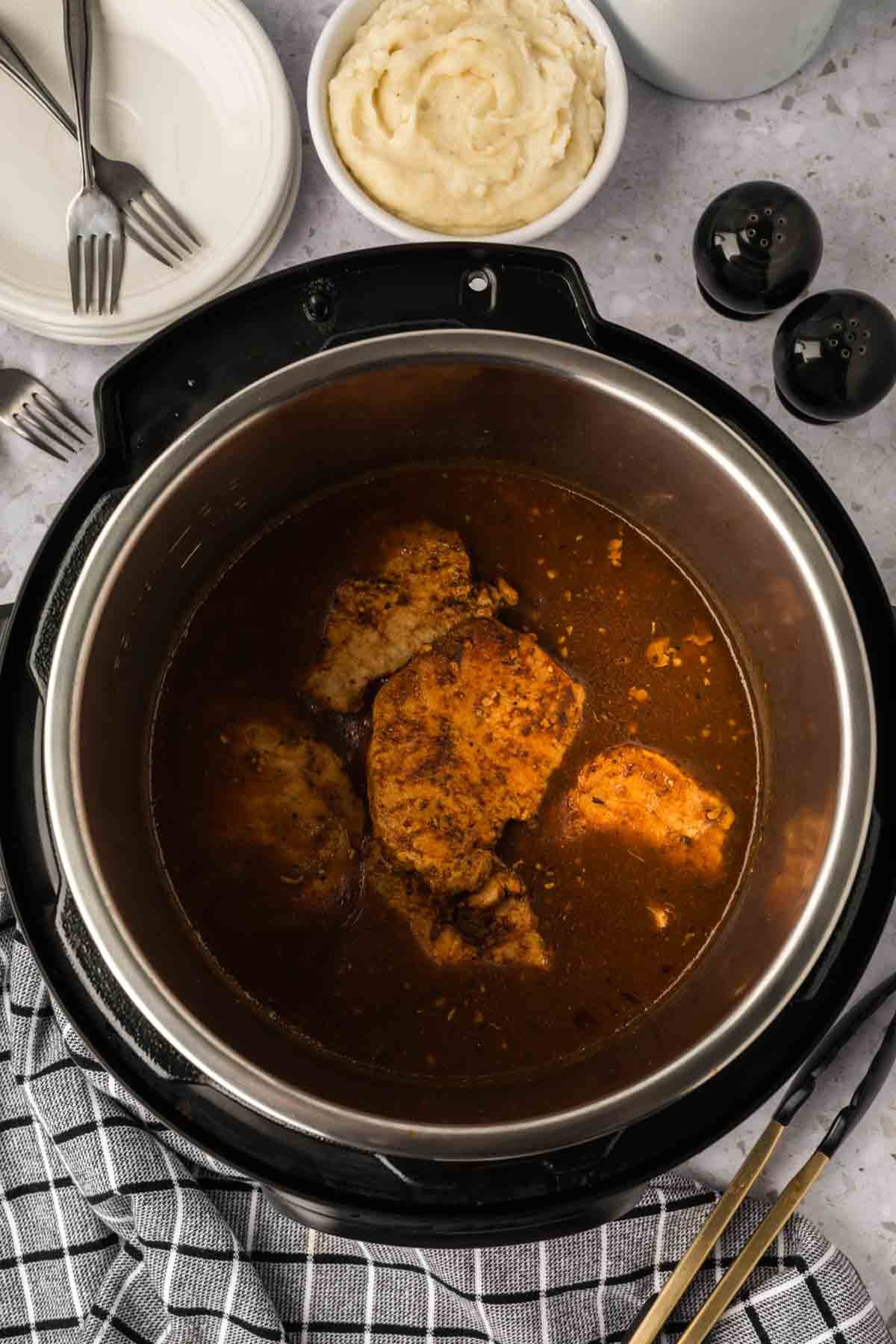 An open Instant Pot reveals tender Instant Pot Pork Chops with Gravy in brown sauce, alongside a bowl of mashed potatoes, stacked plates, forks, salt and pepper shakers, and a gray checked kitchen towel.
