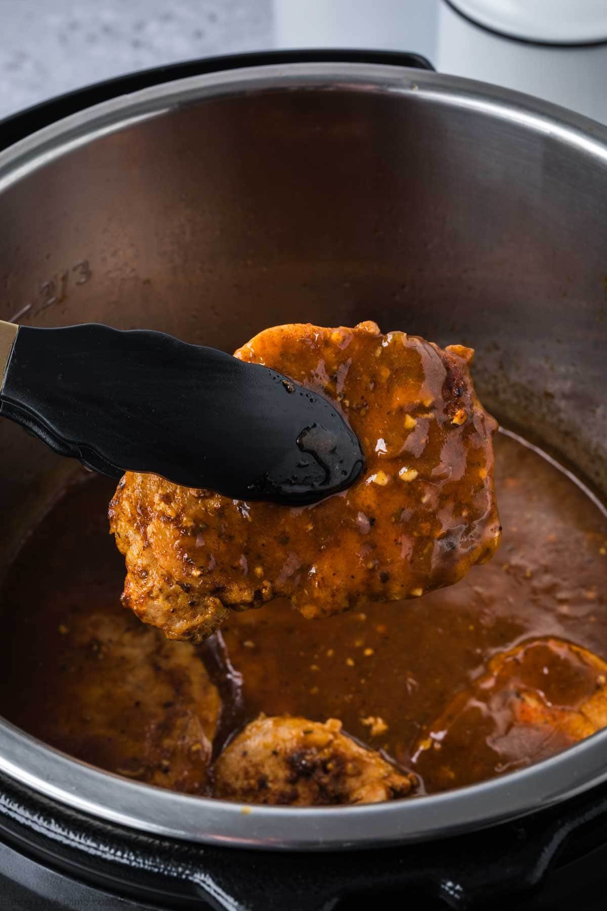 A pair of tongs holds a saucy piece of meat above a pot filled with rich brown gravy and more meat, capturing the savory goodness of Instant Pot Pork Chops with Gravy.