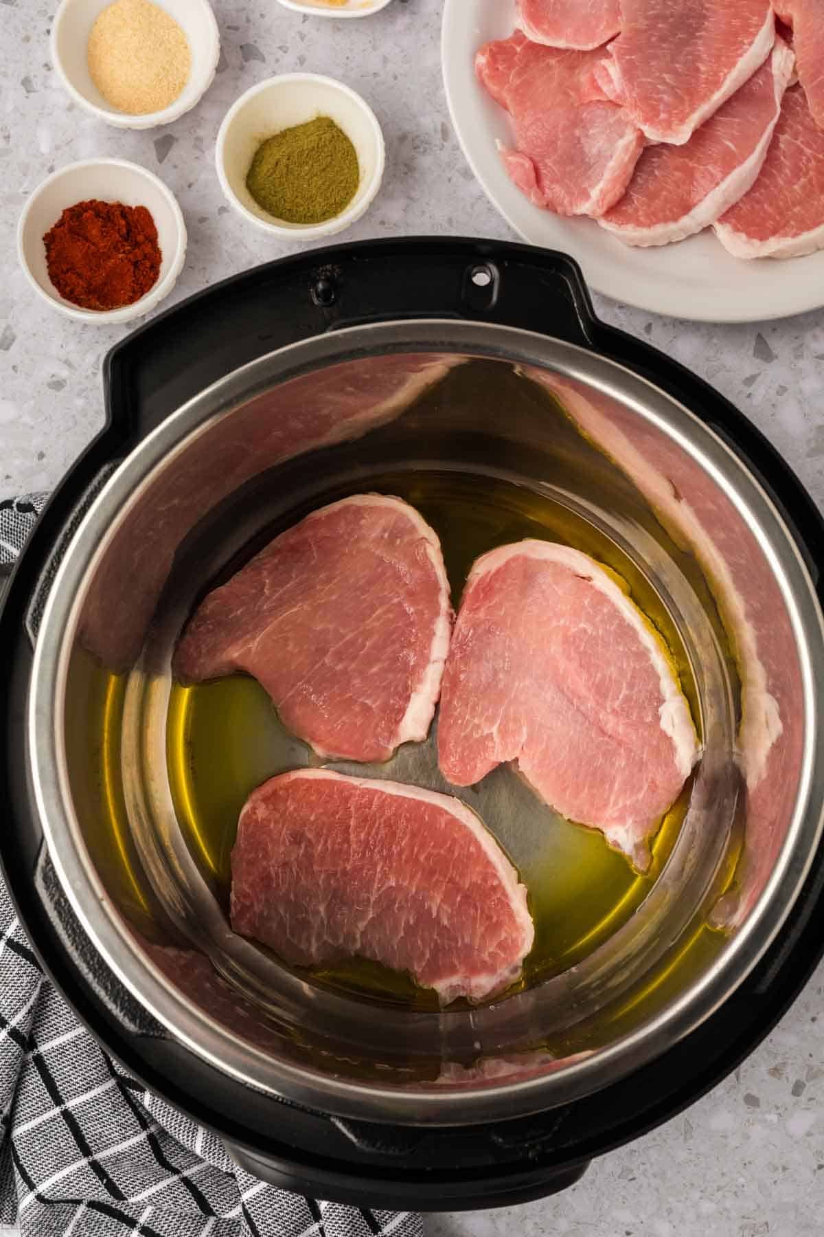Three raw pork chops in oil inside an Instant Pot, ready to become Instant Pot Pork Chops with Gravy, with seasonings in small bowls and extra pork chops on a white plate nearby, all on a gray countertop with a black and white towel.