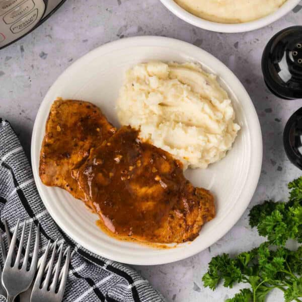 A white plate with Instant Pot Pork Chops with Gravy sits next to creamy mashed potatoes on a gray countertop, surrounded by a black-and-white towel, forks, parsley, and salt and pepper shakers.