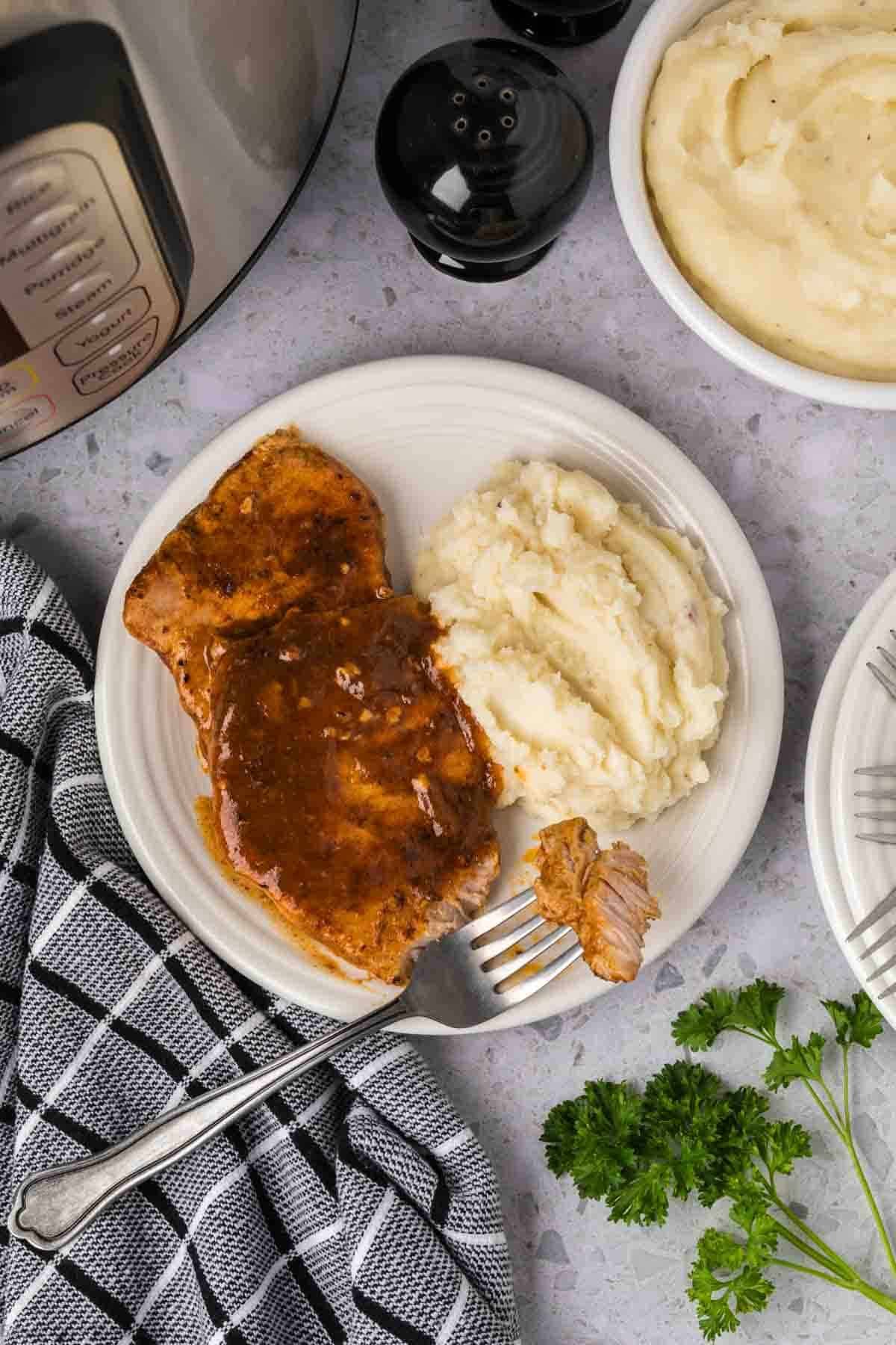 A plate features two pieces of Instant Pot Pork Chops with Gravy beside a scoop of mashed potatoes. A fork rests on the plate, while parsley, a black-and-white towel, and extra mashed potatoes sit nearby.