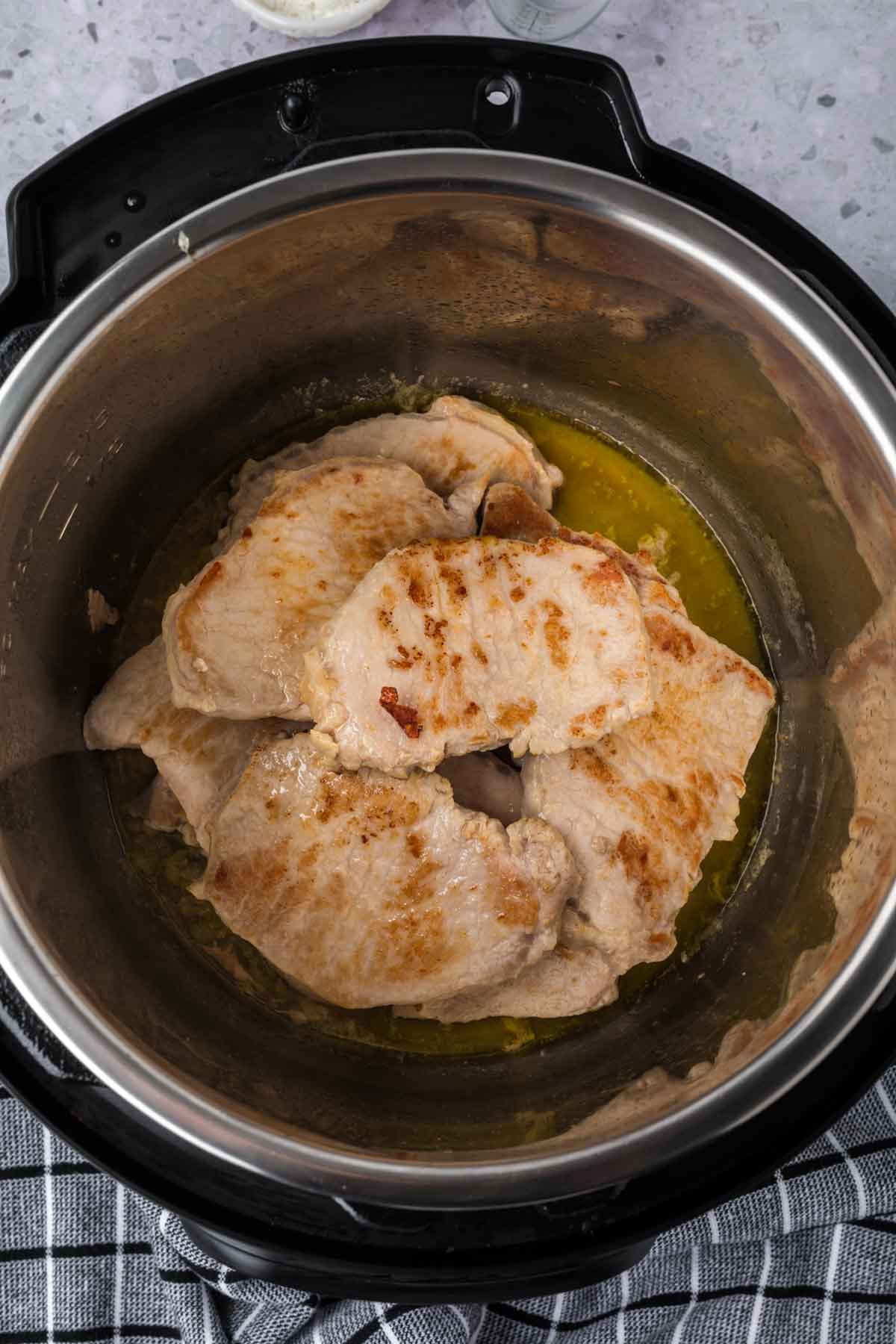 Four browned Instant Pot Pork Chops with Gravy simmer in liquid inside an Instant Pot, viewed from above. The pot rests on a gray checkered cloth atop a speckled countertop.