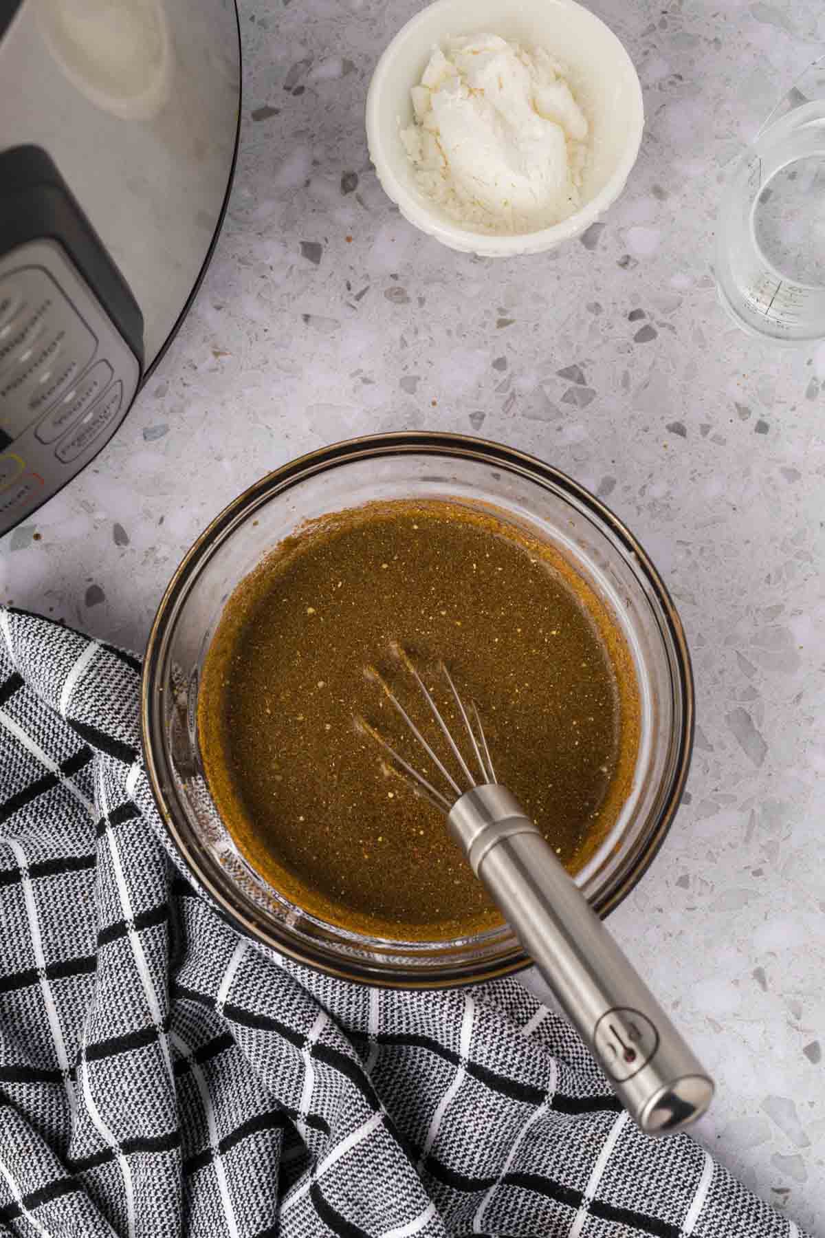 A glass bowl with a dark brown liquid mixture and a whisk sits on a gray countertop next to a checkered towel, a small bowl of white ingredients, and part of an Instant Pot—perfect for making Instant Pot Pork Chops with Gravy.