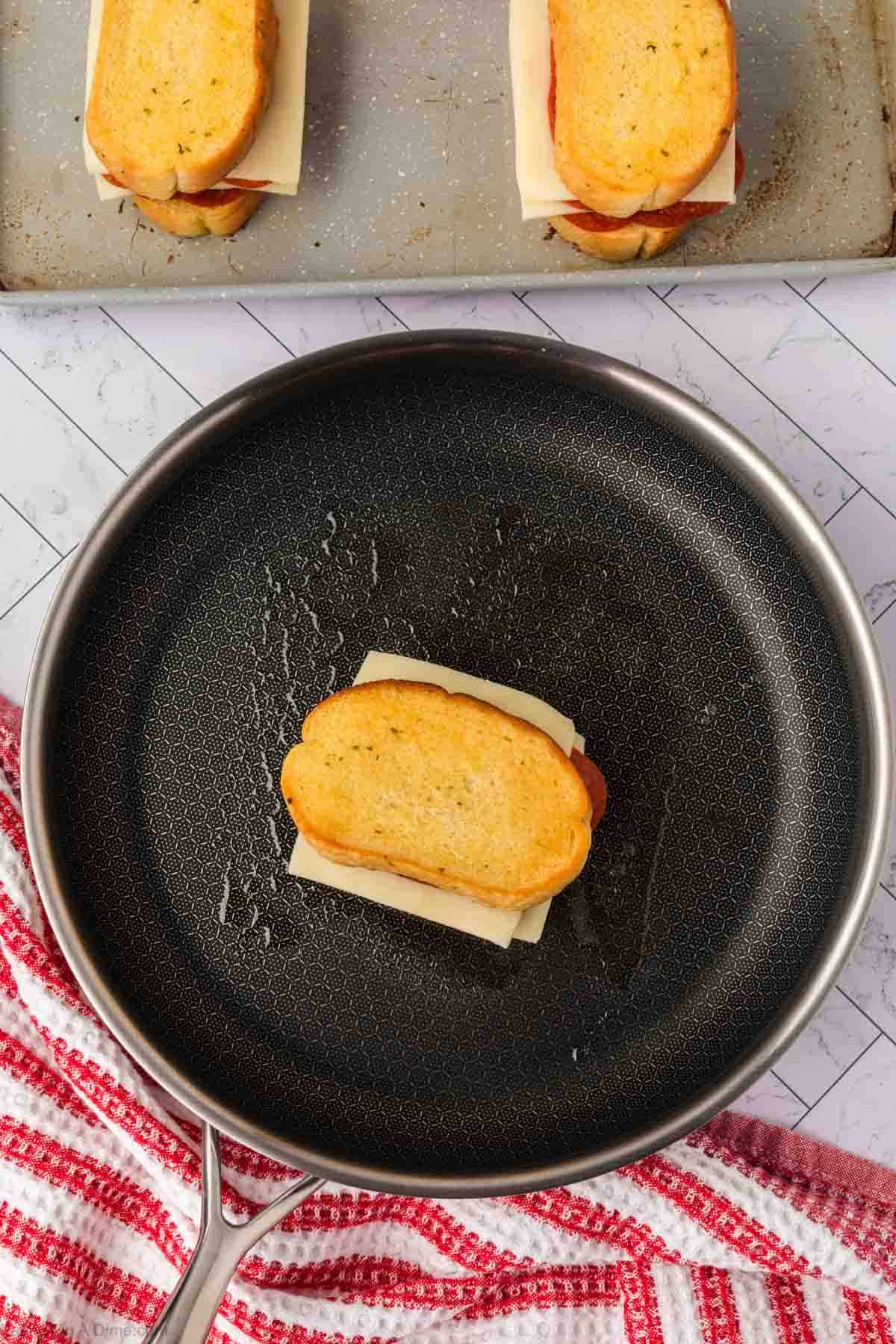 A Pizza Grilled Cheese Sandwich with gooey cheese and garlic bread is sizzling in a frying pan; more sandwiches rest on a baking tray nearby. A red and white striped kitchen towel lies next to the pan.