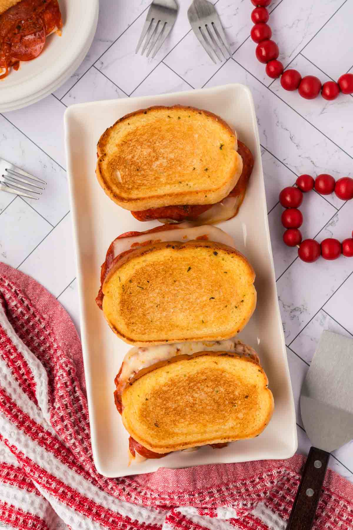 Three toasted Pizza Grilled Cheese Sandwiches with golden-brown bread are arranged in a row on a white rectangular plate. Surrounding the plate are a red and white towel, utensils, red beads, and part of a white plate.