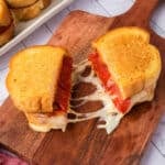 A Pizza Grilled Cheese Sandwich is cut in half on a wooden board, with melted cheese stretching between the halves. The bread is golden and toasted, and slices of pepperoni are visible inside.