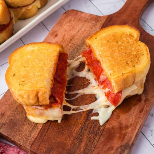 A Pizza Grilled Cheese Sandwich is cut in half on a wooden board, with melted cheese stretching between the halves. The bread is golden and toasted, and slices of pepperoni are visible inside.