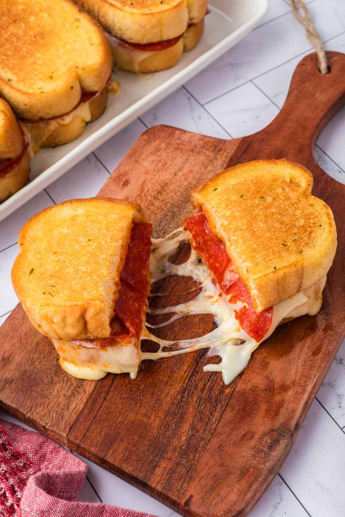 A Pizza Grilled Cheese Sandwich with melted mozzarella and pepperoni is cut in half on a wooden board, gooey cheese stretching between the halves. More sandwiches are stacked on a white tray in the background.