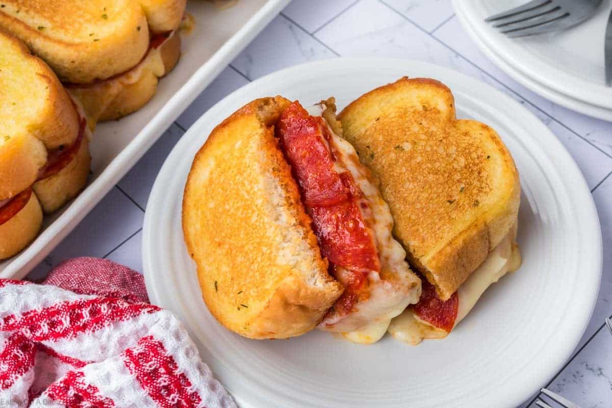 A Pizza Grilled Cheese Sandwich with melted cheese and pepperoni slices is cut in half and served on a white plate, with more sandwiches and a red checkered napkin nearby.