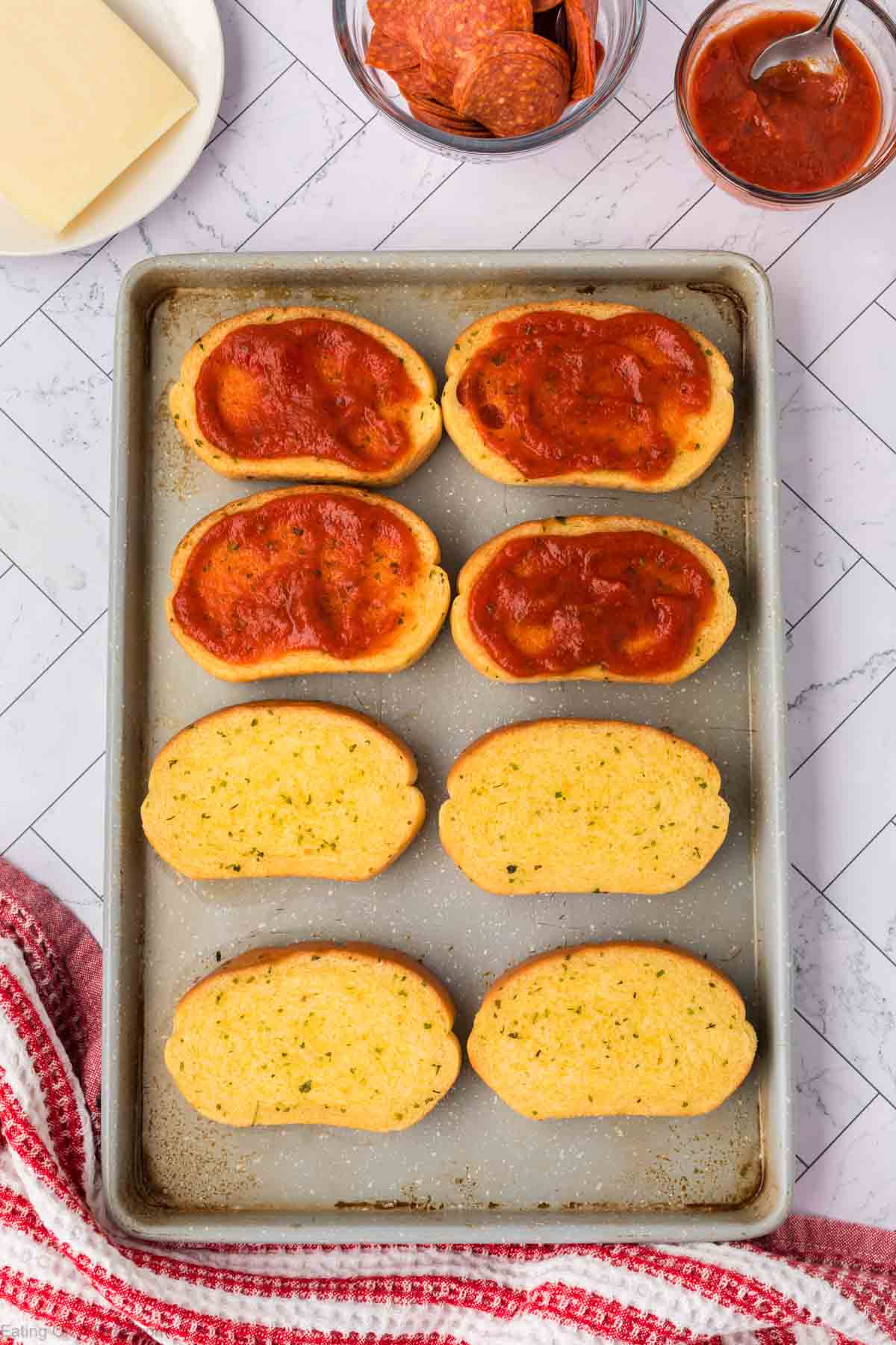 A baking sheet with eight slices of garlic breadโfour topped with pizza sauce, four plainโsurrounded by bowls of pepperoni, pizza sauce, and sliced cheese on a white counter evokes the flavors of a Pizza Grilled Cheese Sandwich.