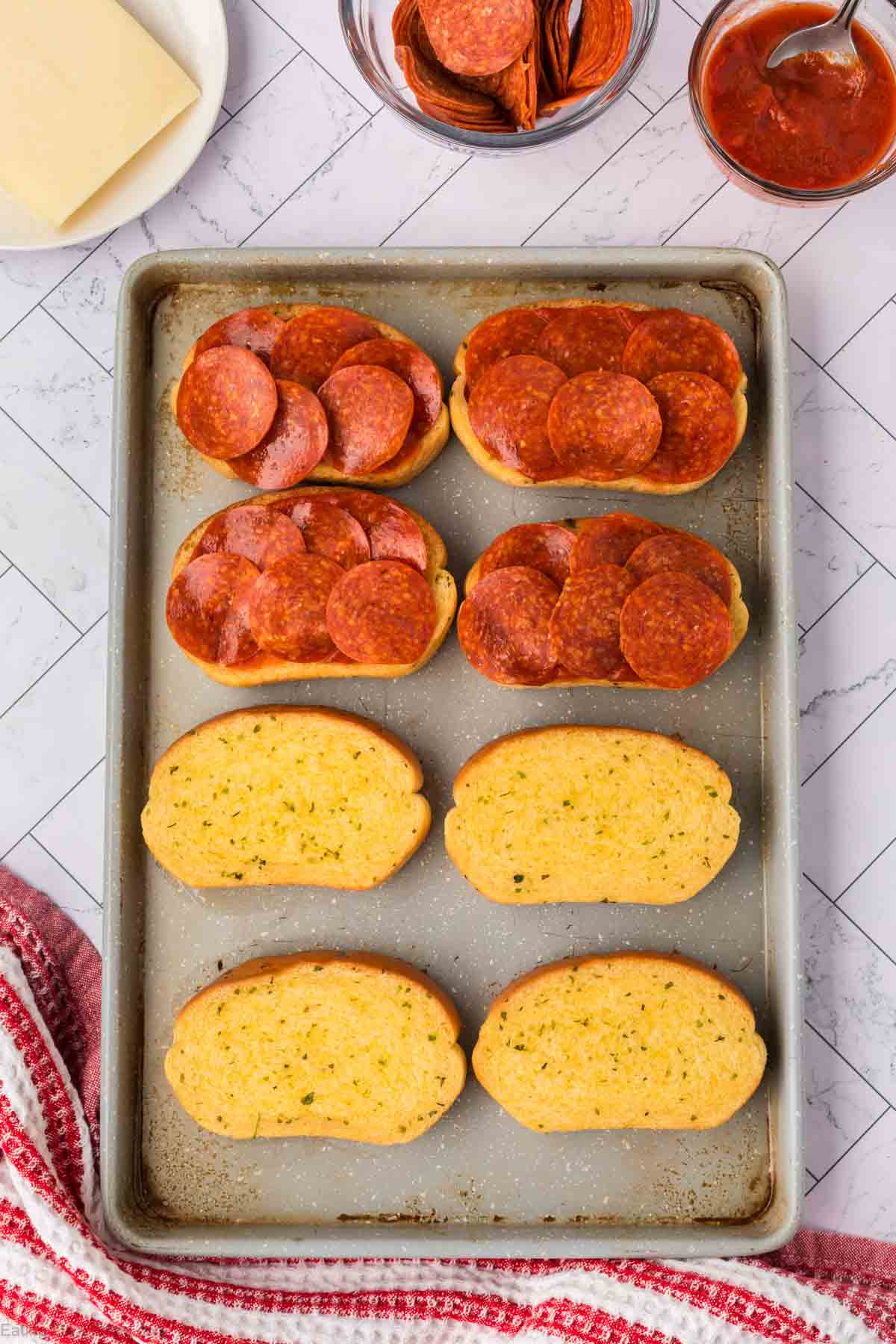 A baking sheet holds six pieces of bread: three topped with tomato sauce and pepperoni slices, like a Pizza Grilled Cheese Sandwich, and three plain. Nearby are bowls of pepperoni, sauce, and a block of cheese on a white tile surface.