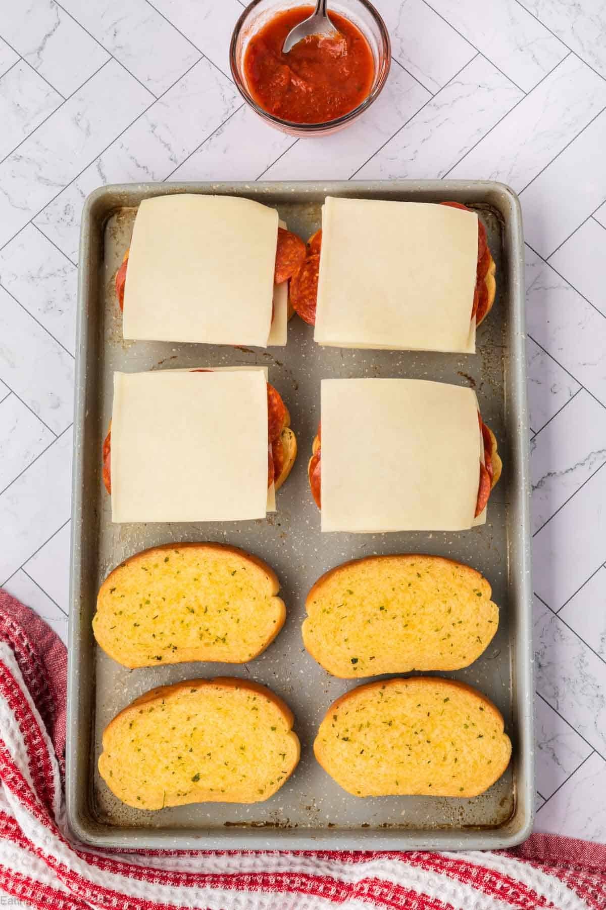 A baking sheet with four open-faced sandwiches: the top row features Pizza Grilled Cheese Sandwich slices with pepperoni and cheese, while the bottom row showcases toasted garlic bread halves. A bowl of red sauce with a spoon sits in the background.
