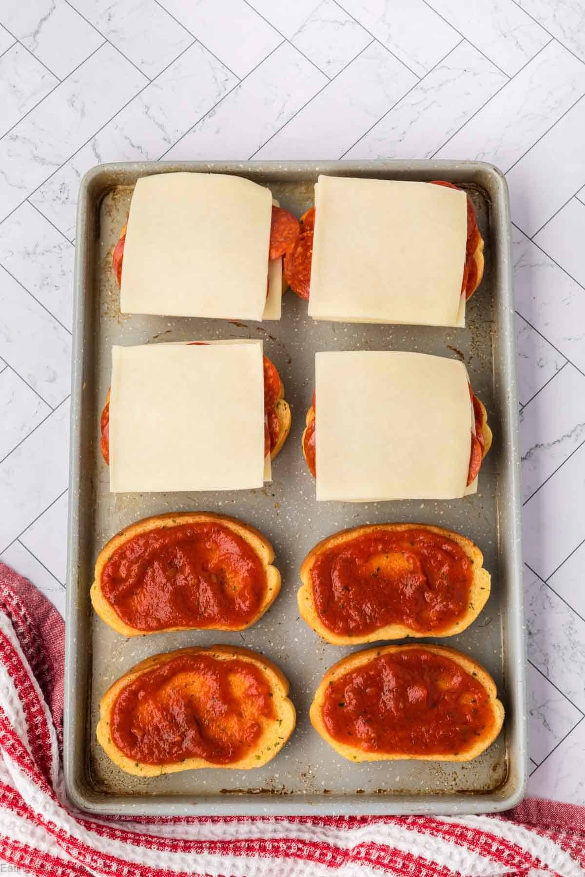 A baking sheet holds six sub rolls: three with pizza sauce, three with pepperoni, other toppings, and cheeseโjust like a Pizza Grilled Cheese Sandwich. A red and white striped towel rests beside the tray on a tiled surface.