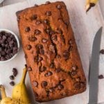 A loaf of chocolate chip banana bread sits on a light wooden surface. Nearby are a serrated knife, a bunch of ripe bananas, and a white bowl filled with extra chocolate chips for the perfect chocolate chip banana bread treat.