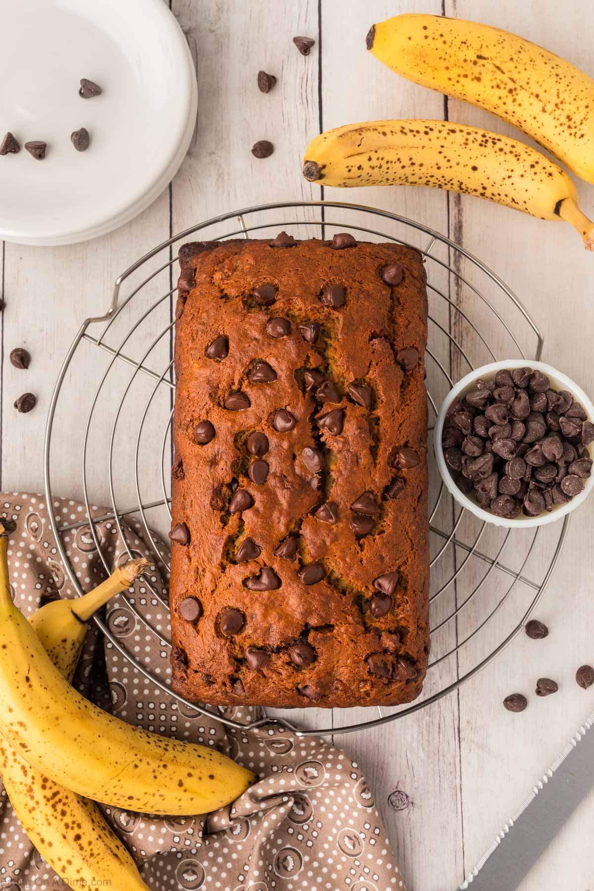 Chocolate Banana Bread loaf on a wire rack with a side of chocolate chips in a bowl and bananas