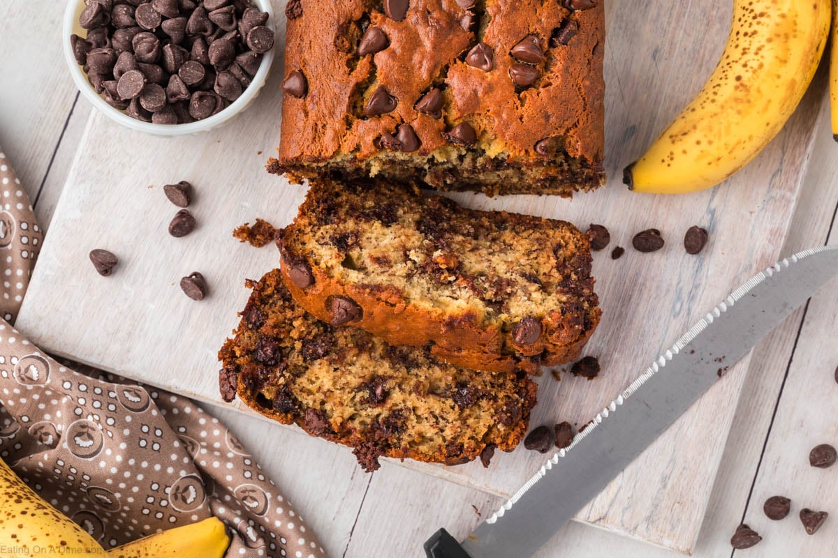 Try this simple and moist Chocolate chip banana bread Recipe! If you love banana bread, then you are going to love this easy chocolate chip banana bread recipe that is made in one bowl! This chocolate chip banana bread is the best! #eatingonadime #bakingrecipes #bananabread Chocolate Chip Banana Bread sliced with a bowl of chocolate chips and bananas nearby.
