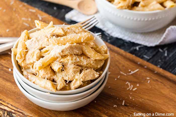 A white bowl full of this Crock Pot Olive Garden Chicken Alfredo Pasta topped with shredded cheese on a wooden board. 
