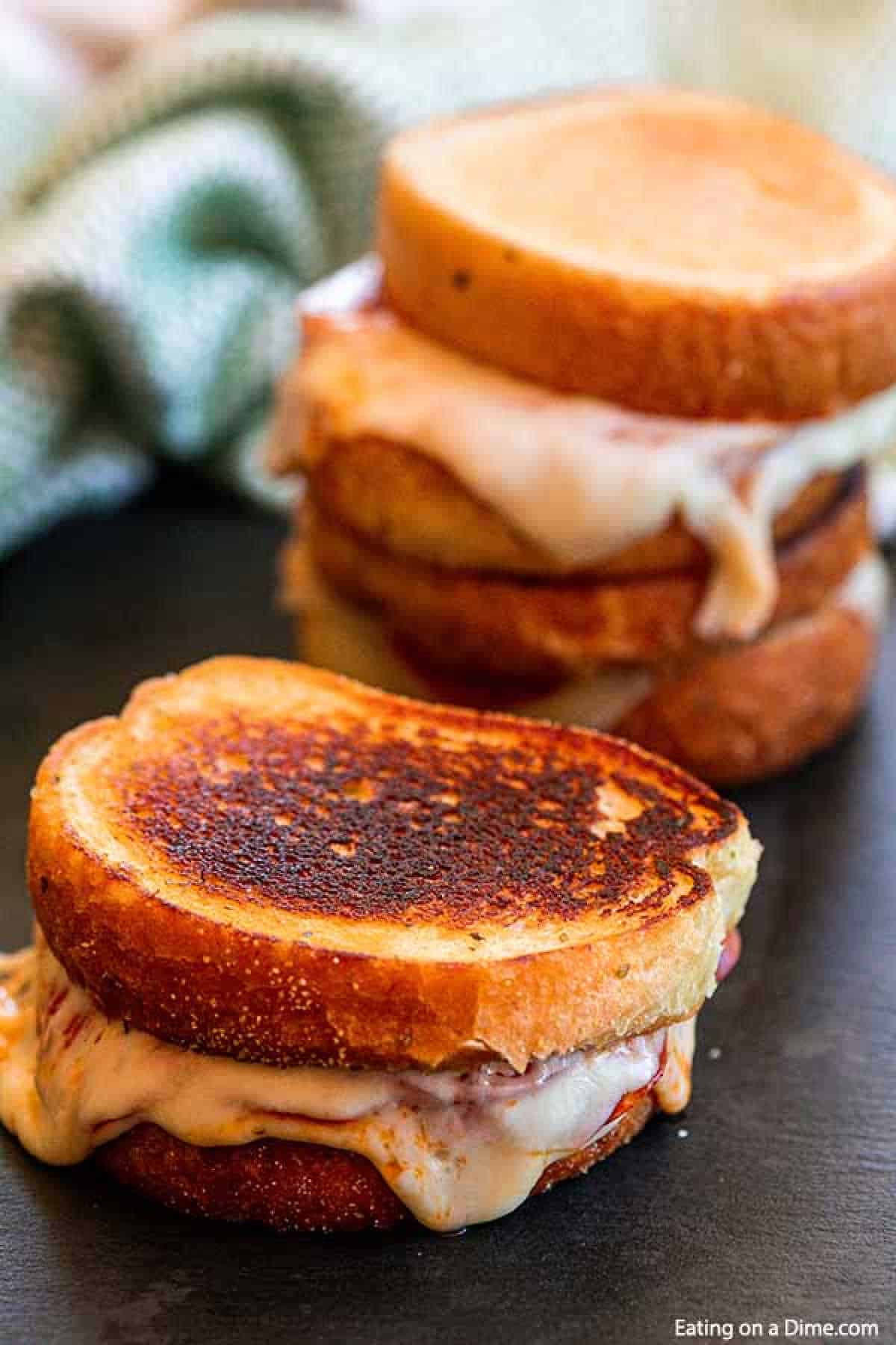 Jazz up plain grilled cheese with this delicious pizza grilled cheese sandwich recipe. Texas Toast makes this super easy. Your kids will go crazy over this! A close-up shot of two pizza grilled cheese sandwiches with golden brown, toasted bread and melted cheese oozing out. A stack of more grilled cheese sandwiches is visible in the background, with a green and white cloth partially visible on a dark surface.