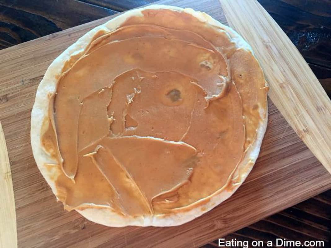 Tortilla on a cutting board with peanut butter spread on the tortilla