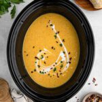 A creamy crockpot butternut squash soup in a black slow cooker, garnished with a swirl of cream, chopped herbs, and red pepper flakes, surrounded by bread, a wooden board, and fresh green parsley.