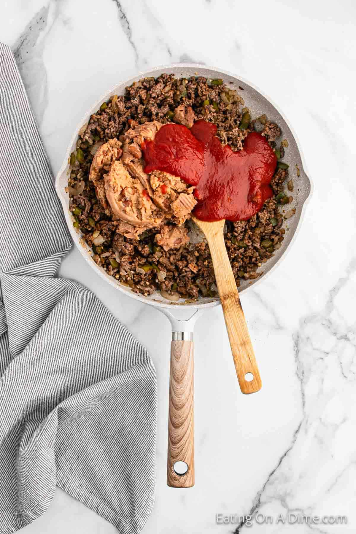 A skillet filled with cooked ground beef, chopped vegetables, and a wooden spoon topped with refried beans and red sauce, ready to be mixed for beef chimichangas. A gray striped dish towel lies next to the skillet on a white marble countertop.