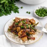A plate of rice topped with Easy Black Pepper Chicken Recipeโstir-fried chicken, red onions, green bell peppers, chopped green onions, and sesame seedsโserved with a fork on the side and fresh parsley and green onions in the background.