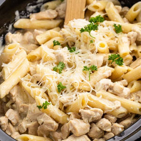A close-up of creamy Crockpot Olive Garden Chicken Pasta with chunks of chicken, topped with grated cheese and parsley, being stirred with a wooden spoon.