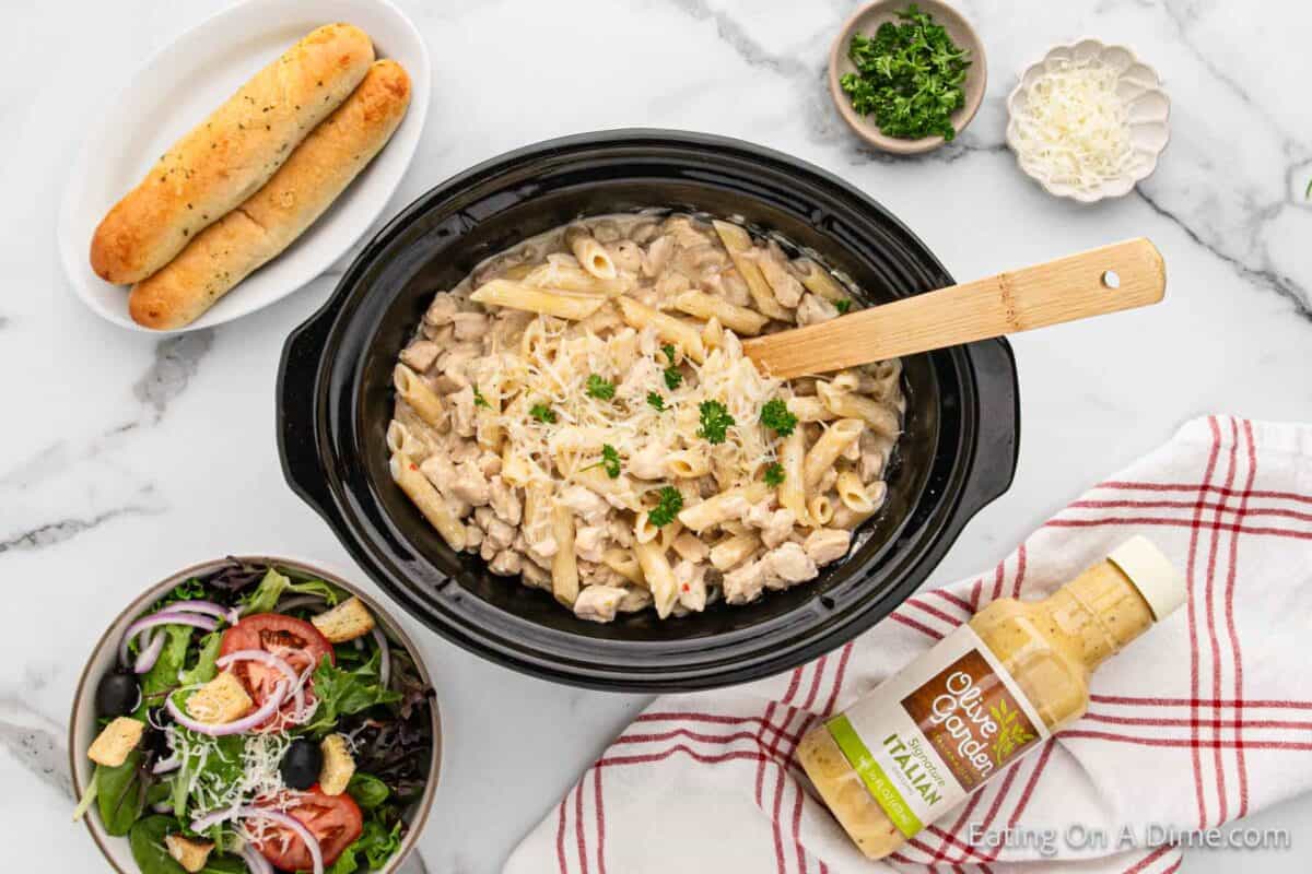 Close up image of chicken alfredo pasta  in the slow cooker with a wooden spoon. A bowl of salad, breadsticks, and salad dressing