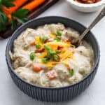 A bowl of creamy chicken pot pie soup, reminiscent of a savory crock pot potato soup with chicken, is garnished with shredded cheese, green onions, and bacon bits on a light-colored surface. A spoon rests inside the bowl while fresh carrots and parsley are visible in the background.