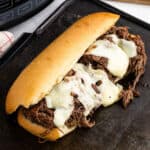 A toasted hoagie roll brimming with shredded beef and melted cheese sits on a dark baking sheet—an irresistible take on classic Shredded Beef Sandwiches, with part of a kitchen appliance visible in the background.