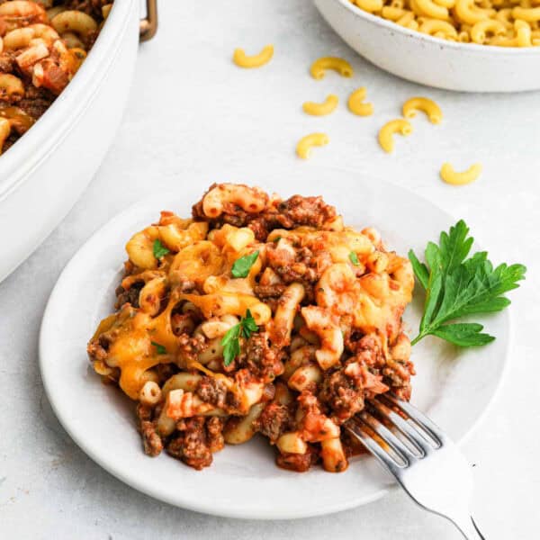A serving of cheesy Hamburger Casserole garnished with parsley sits on a white plate with a fork beside it. A casserole dish, a bowl of macaroni, and scattered pasta surround the plate.