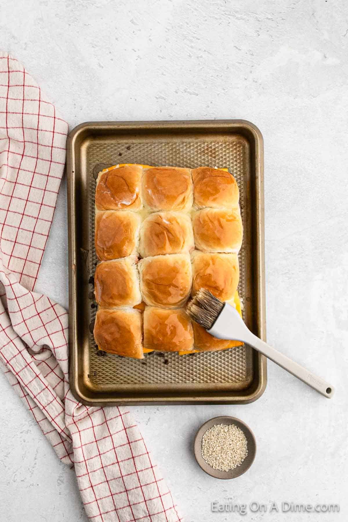 A baking tray with a batch of golden-brown dinner rolls, perfect for a Hawaiian Roll Hamburger Recipe, sits on a light gray surface. A pastry brush rests on the tray, and a small bowl of sesame seeds is nearby. A red and white checkered towel is draped beside the tray.