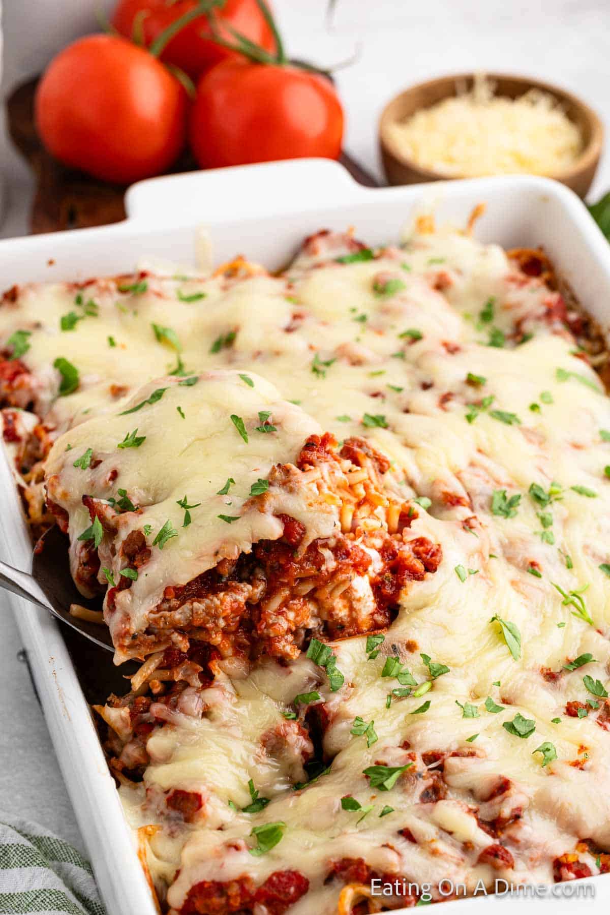 A million dollar spaghetti casserole is shown in a rectangular dish, topped with melted cheese and garnished with chopped parsley. A serving spoon is lifting a portion of the casserole. In the background, you can see fresh tomatoes on a vine and a small bowl of grated cheese.