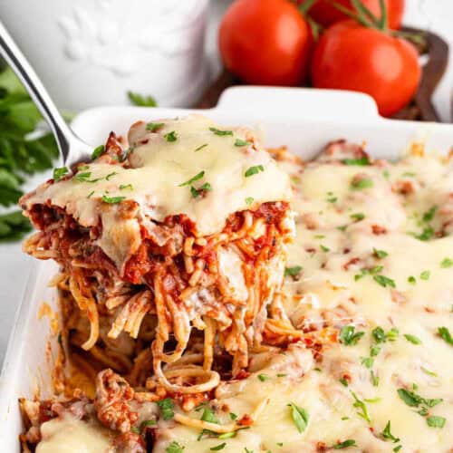 A serving of cheesy baked spaghetti is being lifted from a white casserole dish with a spatula. The million dollar spaghetti recipe features melted cheese and is garnished with chopped parsley. Fresh tomatoes and herbs are visible in the background.