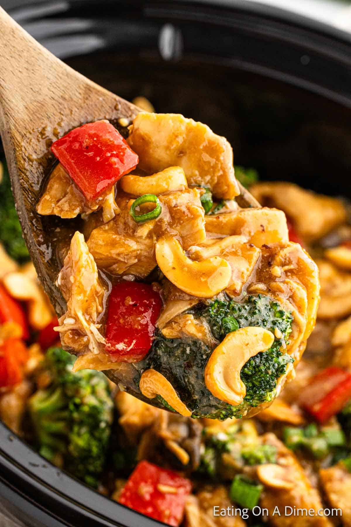 A serving of Cashew chicken with broccoli, cashews and red bell pepper on a wooden spoon with cashew chicken in the background in the slow cooker