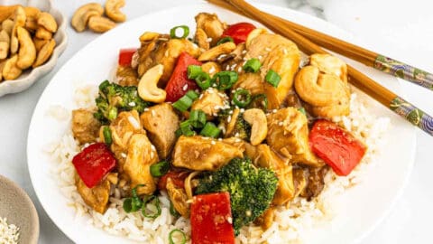 A plate of white rice topped with Crock Pot Cashew Chicken, red bell peppers, broccoli, cashews, and green onions, with chopsticks resting on the plate. Bowls of cashews, green onions, and sesame seeds are nearby.