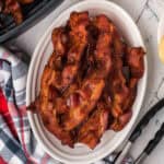 A white oval dish filled with crispy cooked bacon strips, made using an air fryer bacon recipe, sits on a white surface next to metal tongs and a folded plaid kitchen towel.