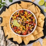 A bowl of vibrant black bean salsa, including corn, black beans, tomatoes, onions, and cilantro, garnished with two lime slices. The bowl is surrounded by a ring of tortilla chips on a round black plate. Lime halves and cilantro leaves are artfully placed in the background.