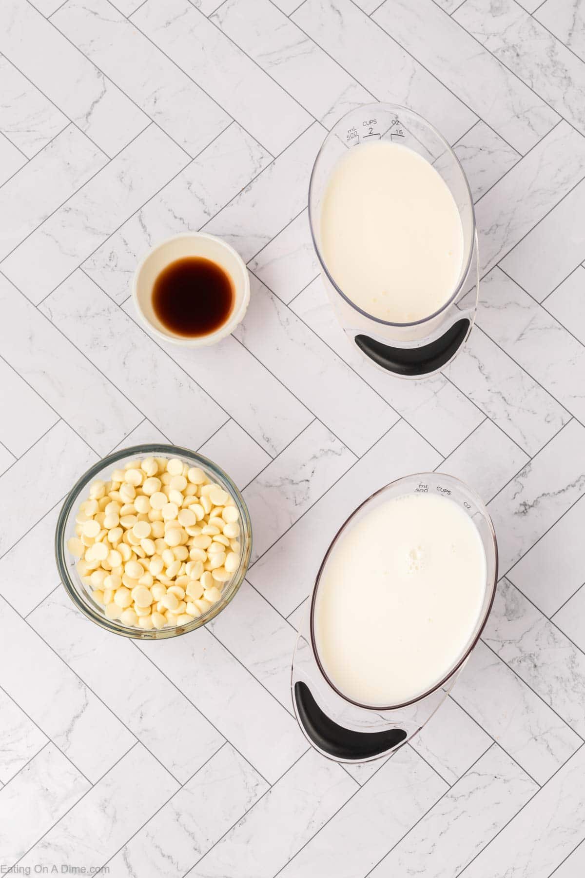 Four ingredients for a crock pot white hot chocolate recipe sit on a white tile surface: vanilla extract in a small bowl, white chocolate chips in a glass bowl, and measuring cups of heavy cream and milk.