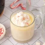 A clear glass mug filled with creamy white hot chocolate, made from a crock pot white hot chocolate recipe, is topped with mini marshmallows and crushed peppermint, sitting on a light tiled surface with empty mugs and candy canes in the background.
