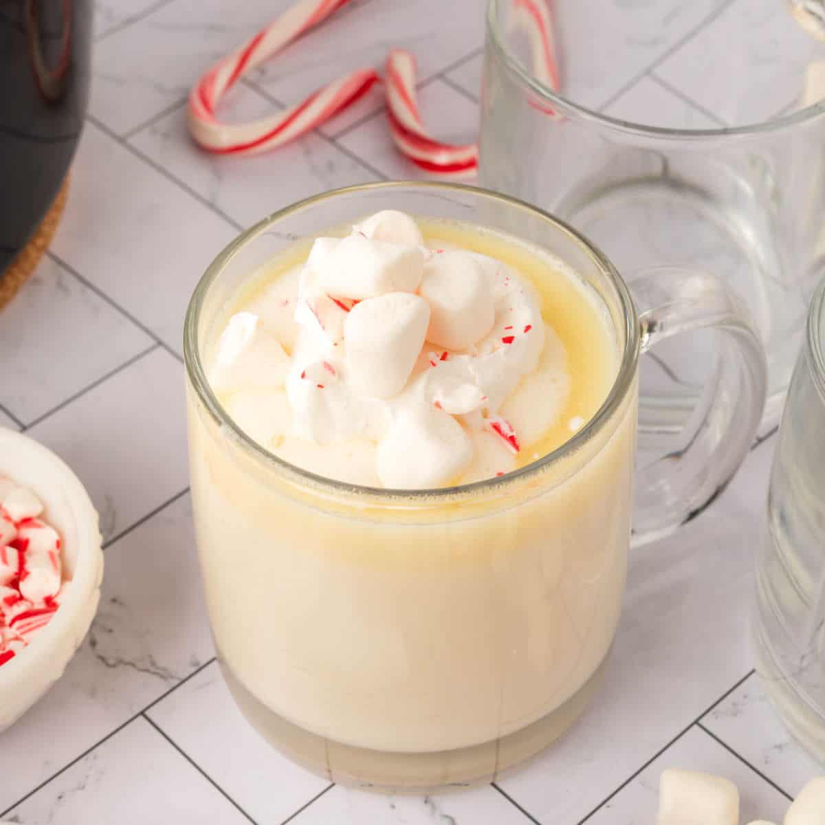 A clear glass mug filled with creamy white hot chocolate, made from a crock pot white hot chocolate recipe, is topped with mini marshmallows and crushed peppermint, sitting on a light tiled surface with empty mugs and candy canes in the background.