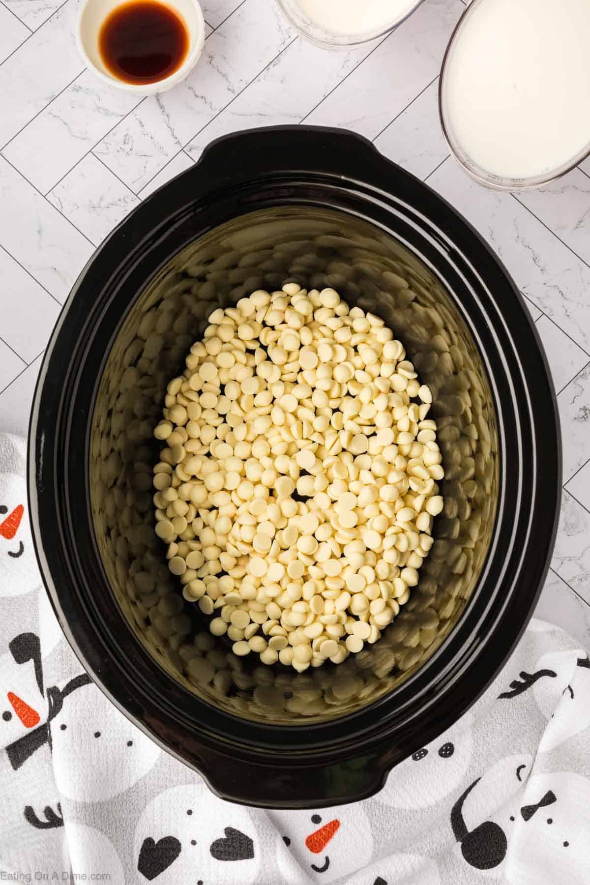 A black slow cooker is filled with white chocolate chips for a cozy crock pot white hot chocolate recipe, surrounded by a cup of vanilla extract, two bowls of milk, and a white towel with snowman and heart patterns.