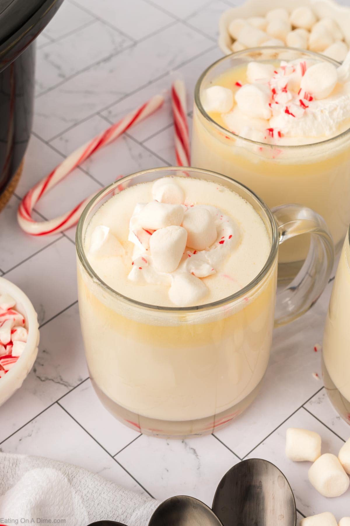 A clear glass mug filled with creamy white hot chocolate, made from a crock pot white hot chocolate recipe, is topped with whipped cream, mini marshmallows, and crushed peppermint. Nearby are extra marshmallows, crushed peppermint, and candy canes on white tiles.